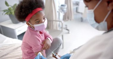 African american female doctor with face mask making injection to girl in hospital room, slow motion. Medicine, healthcare services, childhood and hospital, unaltered.