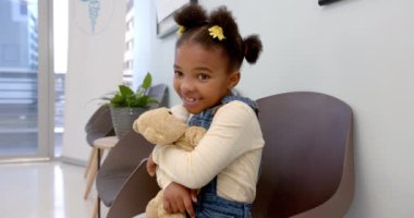 Happy african american girl sitting in hospital waiting room and hugging mascot, slow motion. Medicine, healthcare services, childhood and hospital, unaltered.