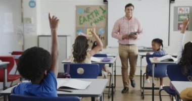 Çeşitli çocuklar el kaldırıyor ve ilkokulda tabletli erkek öğretmen ağır çekimde. Okul, öğrenme, çocukluk, iletişim ve eğitim, değiştirilmemiş.