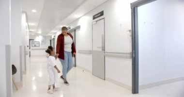 African american mother and daughter in hospital gown holding hands walking in corridor, slow motion. Medicine, healthcare services, family, childhood and hospital, unaltered.