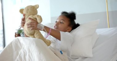 Happy african american girl playing with mascot in hospital bed, slow motion. Medicine, healthcare services, childhood and hospital, unaltered.