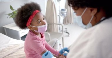African american female doctor with face mask making injection to girl in hospital room, slow motion. Medicine, healthcare services, childhood and hospital, unaltered.
