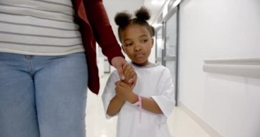 African american mother and daughter in hospital gown holding hands walking in corridor, slow motion. Medicine, healthcare services, family, childhood and hospital, unaltered.