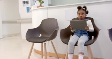 Happy african american girl sitting in hospital waiting room and using tablet, slow motion. Medicine, healthcare services, childhood, communication and hospital, unaltered.