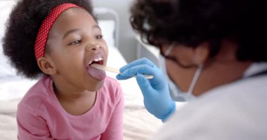 African american female doctor with face mask examining throat of girl in hospital room, slow motion. Medicine, healthcare services, childhood and hospital, unaltered.