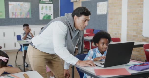 Szczęśliwy Zróżnicowany Nauczyciel Pomaga Chłopcu Przy Biurku Używając Laptopa Podstawówce — Wideo stockowe
