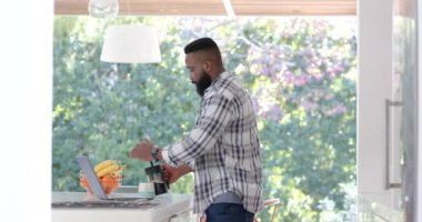Focused african american man using laptop and drinking coffee at home, slow motion. Domestic life, technology, drink, and lifestyle, unaltered.