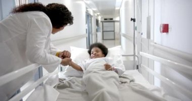 African american female doctor holding hand of girl lying in hospital bed in corridor, slow motion. Medicine, healthcare services, childhood and hospital, unaltered.