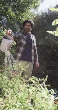 Güneşli bahçede çiçekleri sulayan mutlu melez adamın dikey videosu, yavaş çekim. Yaşam tarzı, doğa, bahçıvanlık ve ev hayatı, değişmemiş..