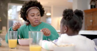 African american mother and daughter eating cereal with milk and talking in kitchen, slow motion. Healthy lifestyle, family, togetherness, food and domestic life, unaltered.
