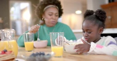 Happy african american mother and daughter eating cereal with milk in kitchen, slow motion. Healthy lifestyle, family, togetherness, food and domestic life, unaltered.