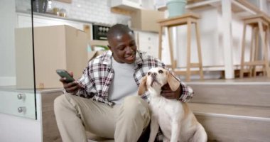 Mutlu Afro-Amerikan adam evde akıllı telefonuyla merdivenlerde oturuyor, evcil köpeğiyle, ağır çekimde. Yaşam tarzı, iletişim, evcil hayvanlar ve ev hayatı, değişmemiş..