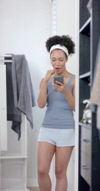 Çift ırklı bir kadının akıllı telefon kullanarak dişlerini fırçaladığı dikey bir video. Yaşam tarzı, iletişim, kişisel bakım ve ev hayatı, değişmemiş..