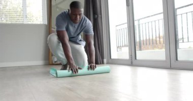 Afrika kökenli Amerikalı bir adam yoga minderini evde yere koyuyor, ağır çekimde. Yaşam tarzı, kişisel bakım, rahatlama ve ev hayatı, değişmemiş..