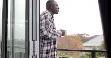 Afrikalı Amerikalı adam balkonda duruyor ve evde kahve içiyor, ağır çekimde. Yaşam tarzı ve ev hayatı, değişmedi..