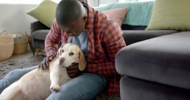 Mutlu Afro-Amerikan adam evinde yerde oturuyor, köpeğini okşuyor ve öpüyor, ağır çekimde. Yaşam tarzı, evcil hayvanlar ve ev hayatı, değişmemiş..