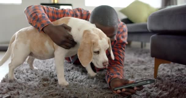 Onnellinen Afrikkalainen Amerikkalainen Mies Makaa Lattialla Käyttäen Älypuhelinta Kotona Lemmikkikoiransa — kuvapankkivideo