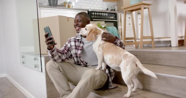 快乐的非洲裔美国人坐在楼梯上使用智能手机 与他的宠物狗在家里 慢动作 生活方式 宠物和家庭生活 — 图库视频影像