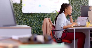 Afrika kökenli Amerikalı sıradan iş kadını laptopunu kullanıyor ve ofiste akıllı telefonla konuşuyor, ağır çekimde. Günlük ofis, iş, iletişim ve iş, değiştirilmemiş.