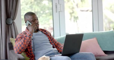 Akıllı telefondaki mutlu Afro-Amerikan adam ve evinde evcil köpeğiyle birlikte dizüstü bilgisayar kullanıyor, ağır çekimde. Yaşam tarzı, iletişim, evcil hayvanlar ve ev hayatı, değişmemiş..