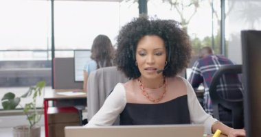 Afrika kökenli Amerikalı kadın telefon setiyle konuşuyor ve ofiste dizüstü bilgisayar kullanıyor. İş, teknoloji ve ofis kavramı