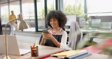 Ofisteki masasında oturan Afro-Amerikan kadın akıllı telefon kullanıyor. İş, teknoloji ve ofis kavramı