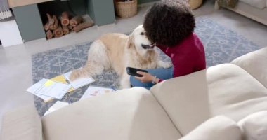 Golden retriever köpeği olan melez bir kadın evde akıllı telefon kullanıyor, ağır çekimde. Yaşam tarzı, hayvan, iletişim ve ev hayatı, değiştirilmemiş..