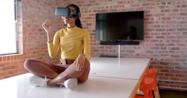 Afrika kökenli Amerikalı iş kadını, VR kulaklık takıyor. Masada oturuyor, ağır çekimde, kopyalama alanında. Günlük ofis, iş, iletişim, teknoloji ve iş, değiştirilmemiş.