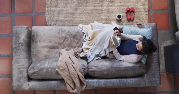 毛布の下にソファーに横たわり 自宅で本を読み ゆっくりと動く女性のオーバーヘッドビュー ライフスタイル フリータイム リラクゼーション 家庭生活 コピースペースなし — ストック動画