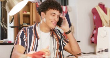 Çift ırklı erkek moda tasarımcısı. Stüdyoda tasarım yapıyor ve akıllı telefondan konuşuyor. Ağır çekimde. Moda, tasarım, yaratıcılık, giyim, teknoloji, iletişim ve küçük işletmeler, değiştirilmemiş.