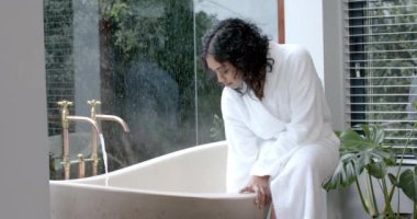 Bornoz giymiş çift ırklı bir kadın küvette oturuyor banyoda banyo yapıyor, ağır çekimde. Yaşam tarzı, rahatlama, kişisel bakım ve ev hayatı, değişmemiş..