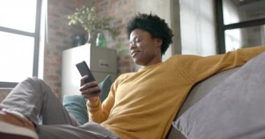 Mutlu Afro-Amerikan adam kanepede oturup akıllı telefon kullanıyor, ağır çekimde. Yaşam tarzı, iletişim ve aile hayatı, değişmedi..