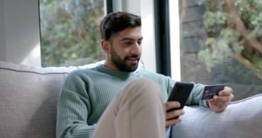 Çift ırklı bir adam evde online alışveriş için akıllı telefon kullanıyor. Ağır çekimde. Yaşam tarzı, iletişim, alışveriş ve ev hayatı, değişmedi..