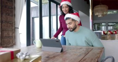 Noel Baba şapkası takan mutlu çift evde video görüşmesi için tablet kullanıyor, ağır çekimde. Yaşam tarzı, kutlamalar, noeller, iletişim ve aile hayatı, değişmemiş.