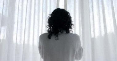 Çift ırklı kadın bornoz giyip perdeleri açıyor ve pencereden eve bakıyor, ağır çekimde. Yaşam tarzı, rahatlama, kişisel bakım ve ev hayatı, değişmemiş..