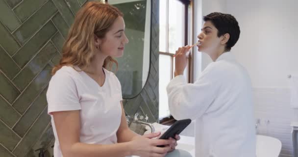 ハッピーな白人レズビアンカップルは一緒に歯を磨き 晴れたバスルームでスマートフォンを使用しています セルフケア トータルネスティ 家庭生活 コミュニケーション — ストック動画