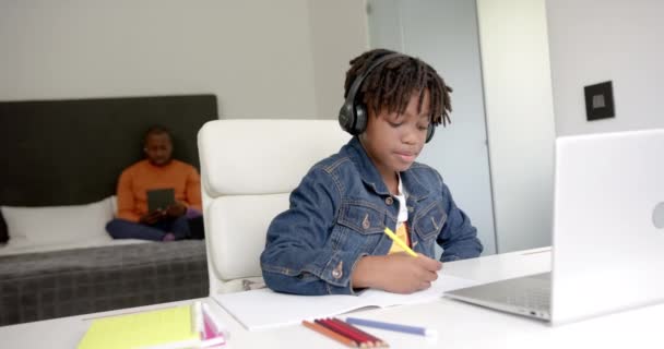 アフリカ系アメリカ人の男の子は 自宅でバックグラウンドで父親とのオンラインレッスンにノートパソコンを使用して ゆっくり動作します 子供時代 コミュニケーション 家庭生活 変わらない — ストック動画