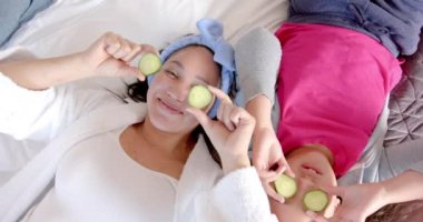 Çift ırklı anne ve kız gözlerinde salatalık dilimleriyle yatakta yatıyorlar. Kişisel bakım, hijyen, aile, annelik, çocukluk, birliktelik ve aile hayatı, değişmemiş..