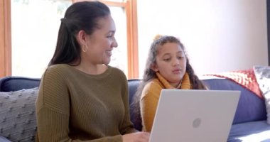 Mutlu çift ırklı anne ve kız kanepeye oturmuş, dizüstü bilgisayar kullanıyor ve güneşli oturma odasında konuşuyor. Aile, annelik, çocukluk, birliktelik ve yaşam tarzı, iletişim ve teknoloji, değiştirilmemiş..