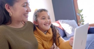 Mutlu çift ırklı anne ve kız kanepeye oturmuş, dizüstü bilgisayar kullanıyor ve güneşli oturma odasında gülümsüyor. Aile, annelik, çocukluk, birliktelik ve yaşam tarzı, iletişim ve teknoloji, değiştirilmemiş..