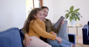 Mutlu çift ırklı anne ve kız kanepeye sarılıp güneşli oturma odasında tablet kullanıyor. Aile, annelik, çocukluk, birliktelik ve yaşam tarzı, iletişim, teknoloji, değişmemiş.