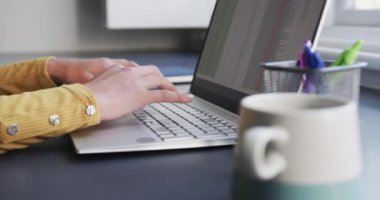 Çift ırklı bir kadının masa başında dizüstü bilgisayarla oturup evde kahve içmesi, ağır çekimde. Uzaktan çalışma, iş, iletişim, yaşam tarzı ve ev hayatı.