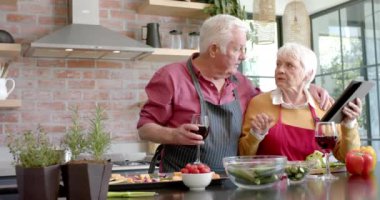 Üst düzey beyaz çift evde mutfakta tablet kullanarak yemek pişiriyor, ağır çekimde. Kıdemli yaşam tarzı, yemek, yemek, tarif, iletişim, sağlıklı yaşam, birliktelik ve aile hayatı, değişmemiş.