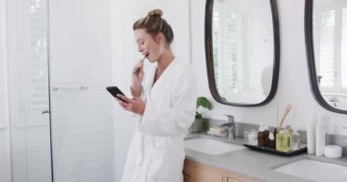 Çift ırklı bir kadın banyoda akıllı telefon kullanarak dişlerini fırçalıyor, ağır çekimde. Kişisel bakım, hijyen, iletişim, yaşam tarzı ve ev hayatı, değişmemiş..