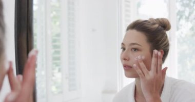 Çift ırklı bir kadın banyodaki aynaya bakarak yüz kremi sürüyor, ağır çekimde. Kişisel bakım, yaşam tarzı ve ev hayatı, değişmedi..