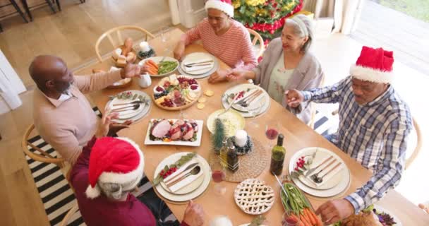 クリスマスの食卓で恵みを語るために手を握っている幸せな多様なシニアの友人 遅い動き クリスマス お祝い そしてシニアライフスタイル 変わらない — ストック動画