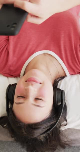 Pionowe Wideo Szczęśliwej Birasowej Kobiety Słuchawkach Leżącej Łóżku Pomocą Smartfona — Wideo stockowe