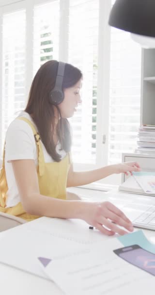 ノートパソコンを使ったヘッドフォンの女性の垂直ビデオ 自宅で働く スローモーション リモートワーキング コミュニケーション ライフスタイル 家庭生活 — ストック動画