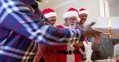 Noel Baba kıyafetli ve Noel şapkalı mutlu kıdemli arkadaşlar evde hediyelerini değiş tokuş ediyorlar, ağır çekimde. Arkadaşlık, Noel, eğlence, gelenek, kutlama, ev hayatı ve yaşlılar yaşam tarzı, değişmemiş.