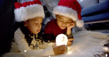Mutlu Afro-Amerikan kardeşler Noel şapkası takmış kar küresiyle oynuyorlar, ağır çekimde. Çocukluk, Noel, kutlama, gelenek, aile, birliktelik ve aile hayatı, değiştirilmemiş.